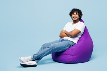 Full body young happy Indian man he wear white t-shirt casual clothes sit in bag chair hold hands crossed folded isolated on plain pastel light blue cyan background studio portrait. Lifestyle concept.