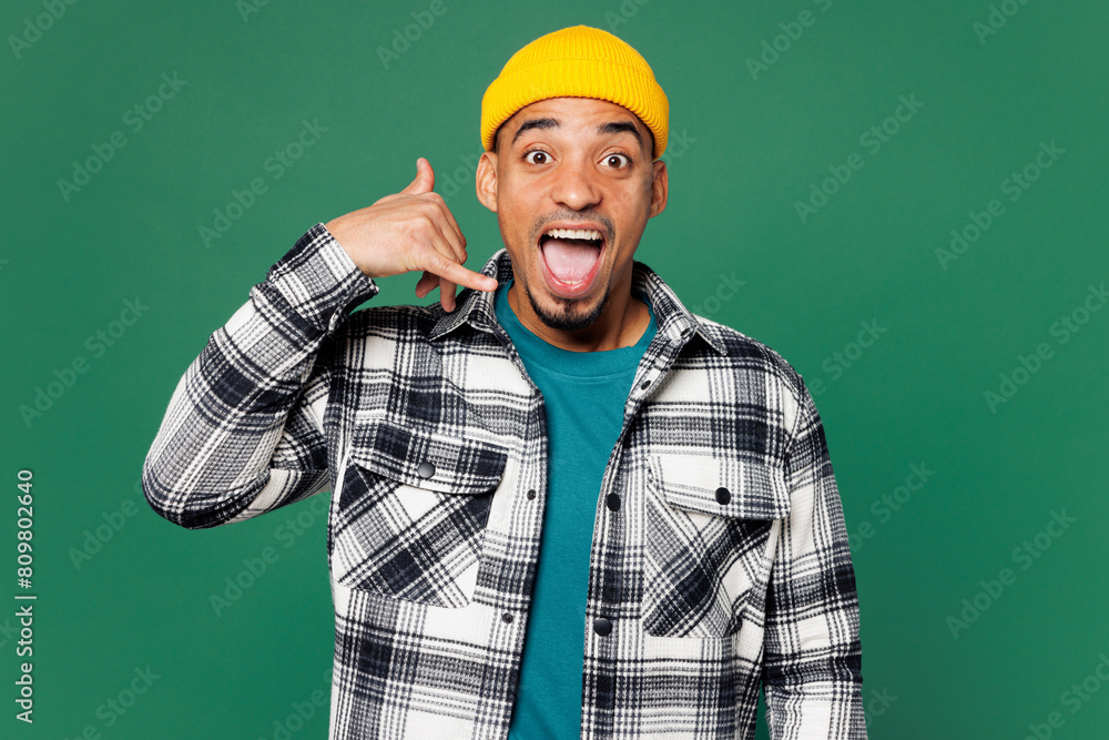 Wall mural Young surprised shocked man of African American ethnicity he wear shirt blue t-shirt yellow hat doing phone gesture like says call me back isolated on plain green background studio. Lifestyle concept. - Wall murals