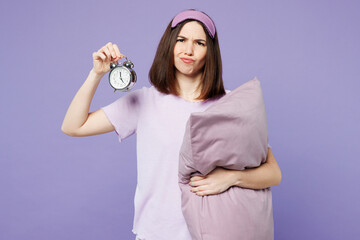 Young sad woman wear pyjamas jam sleep eye mask rest relax at home hold pillow show clock alarm...