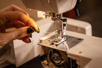 A close-up of a girl's hand with a black manicure feeds a thread into a needle of a sewing machine.