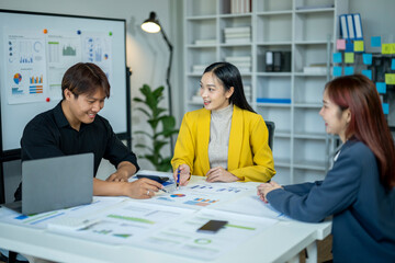 Three people are sitting at a table with papers and a laptop. They are discussing something and seem to be in a professional setting