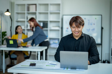 A man is sitting at a desk with a laptop in front of him. He is smiling and he is enjoying his work. The scene takes place in an office setting with a whiteboard and several books on shelves
