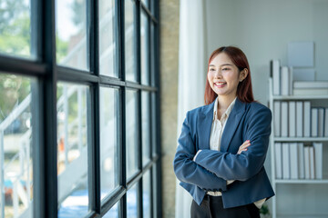 A woman in a blue jacket and white shirt stands in front of a window with her arms crossed. She has a smile on her face and she is confident and professional