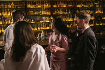 Fototapeta premium Men meet brunette women in nightclub and invite drink cocktails. Friends pay attention to bar counter with elite alcohol in club