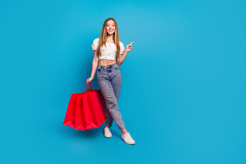 Full size photo of nice young girl hold bags point finger empty space wear top isolated on blue...