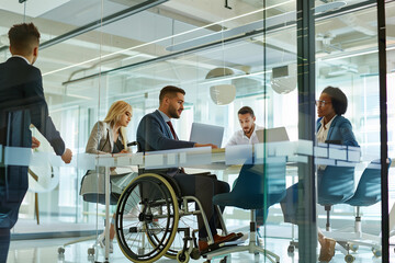 In the bright, open atmosphere of a glass-walled office, a young and diverse business team, led by a businessman in a wheelchair, conducts a productive meeting with laptops in hand