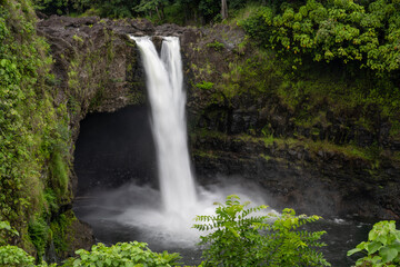 Fototapeta premium Waterfall in the rainforest, the soft, long speed of the water flowing down to the river on a mountain cave background, surrounded by green grass and trees. RAINBOW FALLS Big Island Hawaii USA