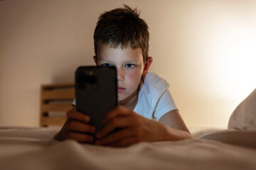Child playing with mobile phone, lying on a bed. Technology and internet concept. Boy laying on bed...