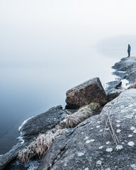 A lone person stands by the edge of a serene, mist-covered lake. The early morning atmosphere is...