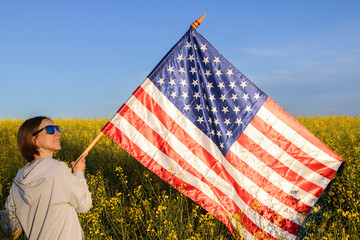 woman holds a large American flag against background of blue sky and rapeseed field on sunny day....