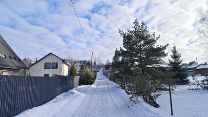 In winter, snow lies on the ground, trees and roofs of houses. Along the streets there are metal and wooden fences and power line poles. There are tire tracks on the snow-covered road. Sunny weather