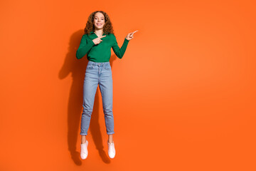 Full length photo of excited cute lady dressed green shirt jumping high pointing two fingers emtpy space isolated orange color background