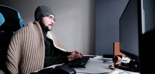 Banner. Young guy in glasses and a warm hat, wrapped in a blanket, works at a computer. It's cold in the room. The concept of turning off heating and gas, lowering the temperature in the room