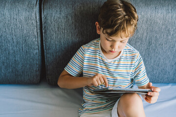 A young boy is sitting on a gray couch, deeply focused on playing tablet. He is wearing a t-shirt...