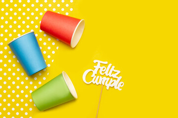Cartel de feliz cumpleaños junto a unos vasos de cartón de colores sobre un fondo amarillo. Vista...