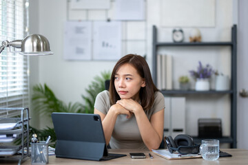 A woman is sitting at a desk with a laptop and a tablet. She is looking at the laptop screen and...