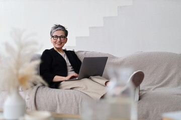 A woman sits comfortably on the sofa, using her laptop for leisure and communication at home.