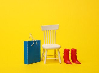 Miniature chair with a shopping bag and doll boots, yellow background. Shopping, sale concept