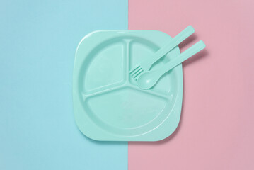 Separate plastic plate with spoon and fork for feeding baby on pink blue background. Top view