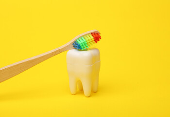 Plastic tooth model with rainbow toothbrush on a yellow background. Dental care