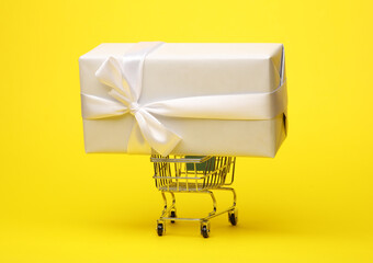 White gift box with bow in mini shopping cart on yellow background