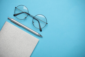Notepad with silver cover and pen with eyeglasses on blue background. Flat lay