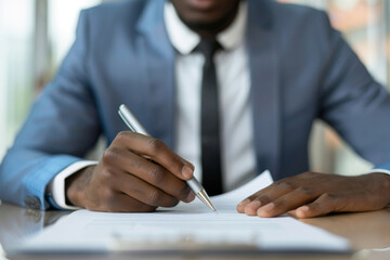 Close-up of Afro-American businessmen signing a formal multicultural business agreement document with diverse people in the office. Showcasing the official corporate paperwork