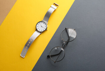 Stylish wristwatch with eyeglasses on yelow gray background. Women's accessories. Top view