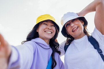Two young attractive multiethnic women take selfies and laugh outdoors. Smiling female friends of different races in casual clothes and summer hats have fun and take pictures in nature.