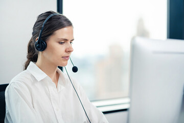A woman wearing a headset is sitting at a desk in front of a computer monitor. She is focused on...