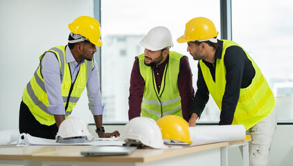 Three men wearing safety gear and hard hats are standing around a table with blueprints. They are...