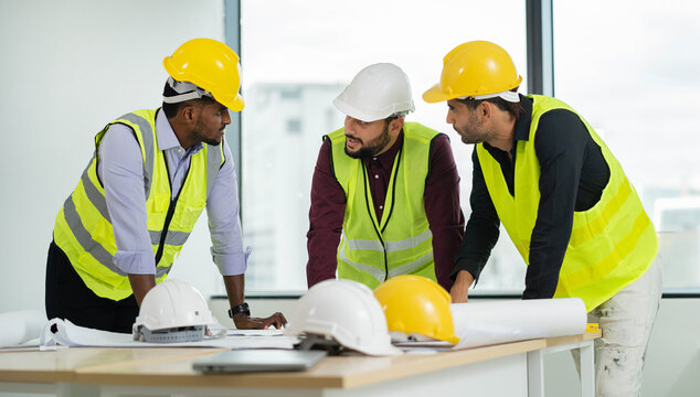 Three men wearing safety gear and hard hats are standing around a table with blueprints. They are discussing the plans and making sure everything is in order. Scene is serious and focused