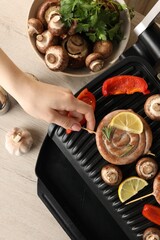 Woman cooking homemade sausages with mushrooms and bell pepper on electric grill at wooden table, top view