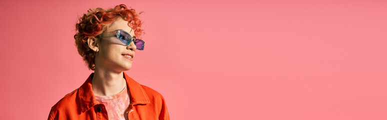 A stylish woman with red hair wearing sunglasses against a bright pink background.