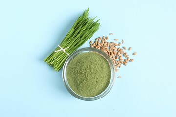 Wheat grass powder in glass bowl, seeds and fresh sprouts on light blue table, flat lay