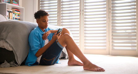 Anxious Teenage Boy Sitting On Bedroom Floor At Home With Mobile Phone Concerned About Social Media