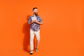 Full length photo of cool guy with red beard wear print shirt hold tablet look at offer empty space isolated on orange color background