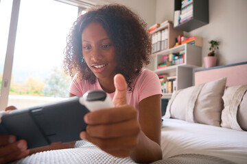 Teenage Girl Lying On Bed At Home Playing With Handheld Gaming Device