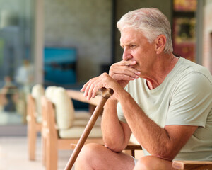Worried Senior Man Holding Walking Stick Sitting In Chair At Home 