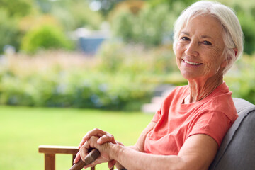 Portrait Of Senior Woman Holding Walking Stick Sitting In Chair At Home Looking Out At Garden