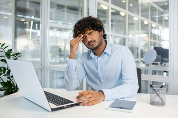 A young man in a blue shirt feels a headache while working on his laptop in a modern office...