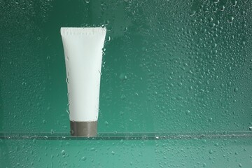 Tube with moisturizing cream on green background, view through wet glass. Space for text