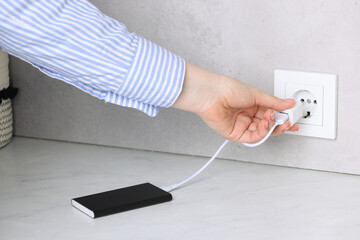 Woman plugging power bank into socket at white table indoors, closeup