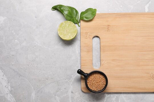 Wooden cutting board, lime, basil and dry mustard on grey marble table, flat lay. Space for text