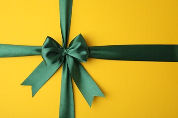 Green satin ribbon with bow on yellow background, top view