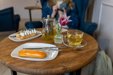 A casual cafÃ© setting with a wooden table featuring an eclair on a white plate, accompanied by silver cutlery and a glass cup. A teapot and a purple tulip in a glass vase add to the ambiance