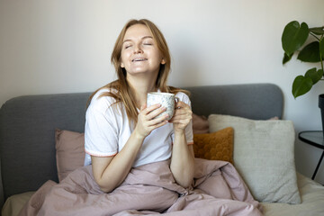 Portrait of a pretty young blond girl holding a mug with hot drink. Woman relaxing on cozy bed at home. Drinks and leisure concept