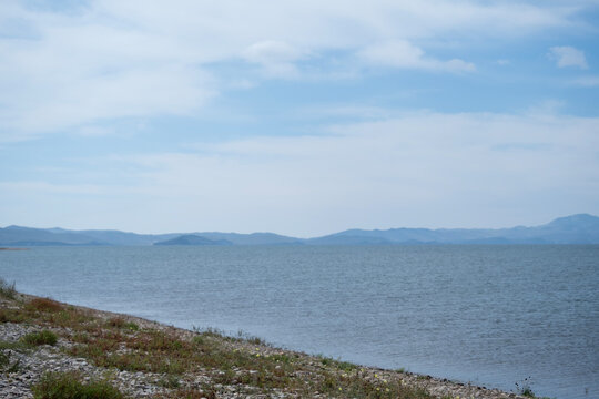 Water landscape, distant view of Lake Baikal from land in corner covered with rocks and wild grass, on horizon faintly visible mountains of opposite shore, summer day with white blue clouds in sky