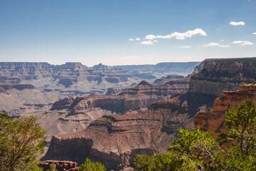 Aerial view of Grand Canyon, Arizona, under clear blue sky. Red rock formations, green shrubs, geological layers, sunlight shadows, rugged texture, hazy horizon. Iconic American landmark.