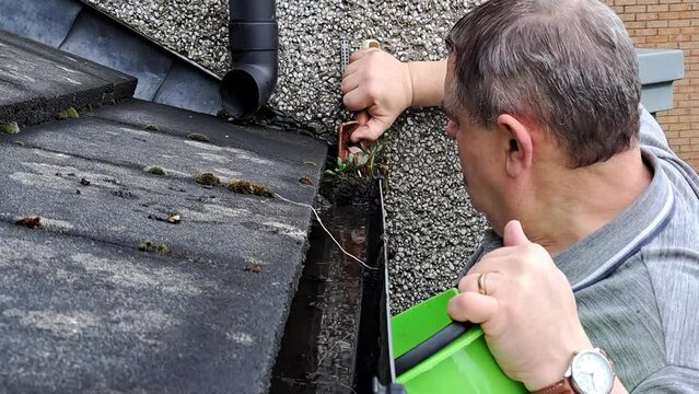 Man with green bucket on ladder cleans dirt and weeds from gutter on tiled roof